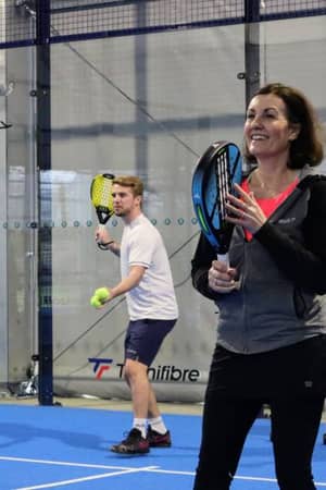A smiling woman with a padel racket in an indoor court, coaching session under the padel4all brand, man in the background preparing to play.