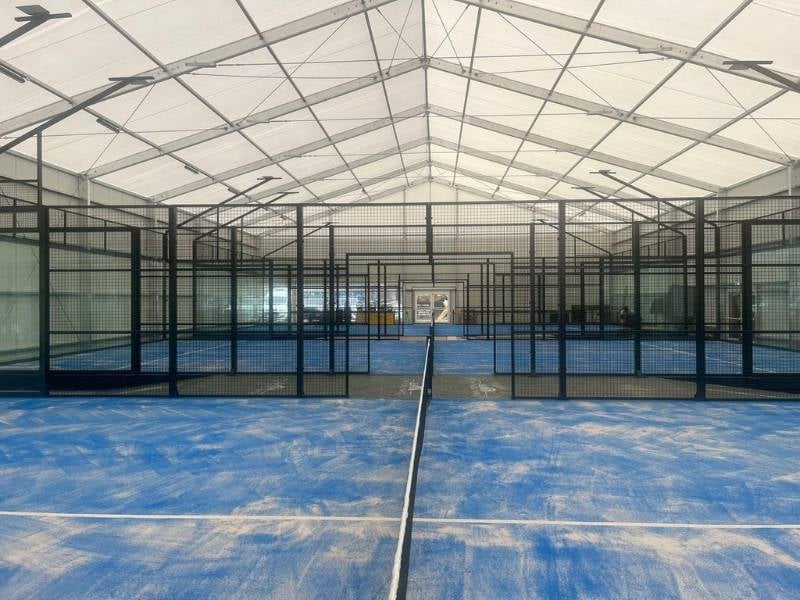 Indoor padel facility with multiple blue courts and black mesh partitions under a white canopy.