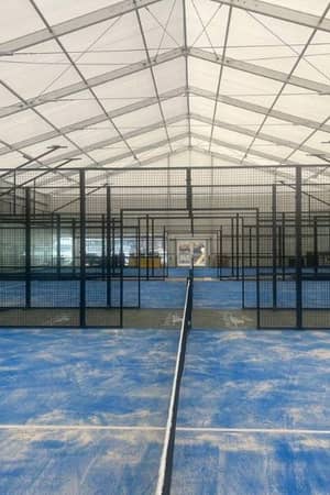 Indoor padel facility with multiple blue courts and black mesh partitions under a white canopy.