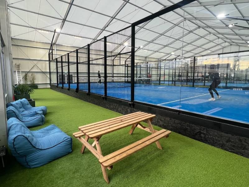 Indoor padel courts with surrounding glass walls, players in action, bean bag seating, and a picnic table on artificial grass.