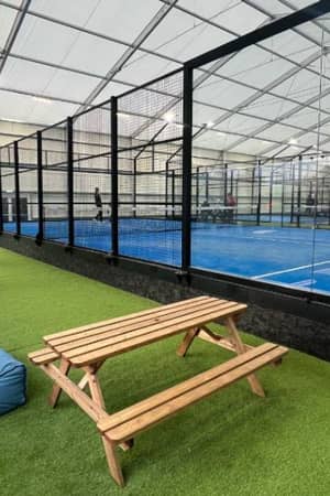 Indoor padel courts with surrounding glass walls, players in action, bean bag seating, and a picnic table on artificial grass.