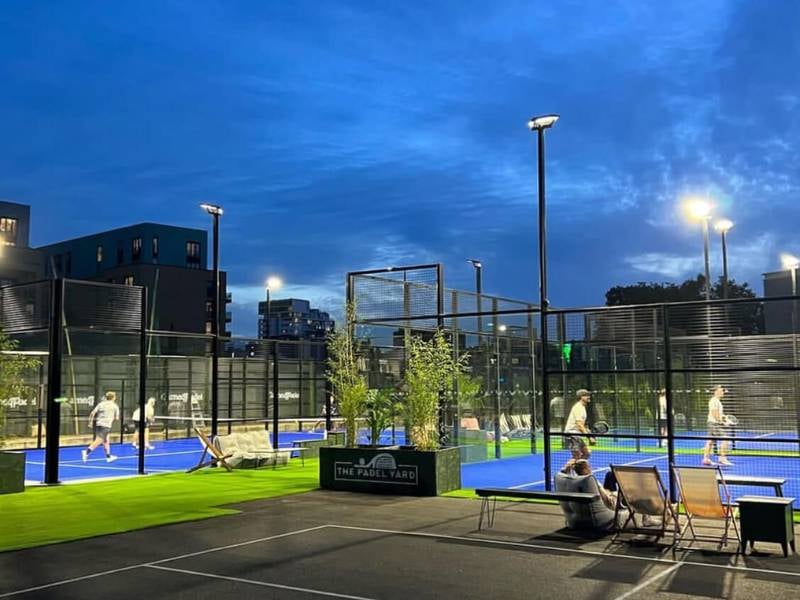 Evening view of The Padel Yard's outdoor courts with players and an urban backdrop, illuminated by lights.
