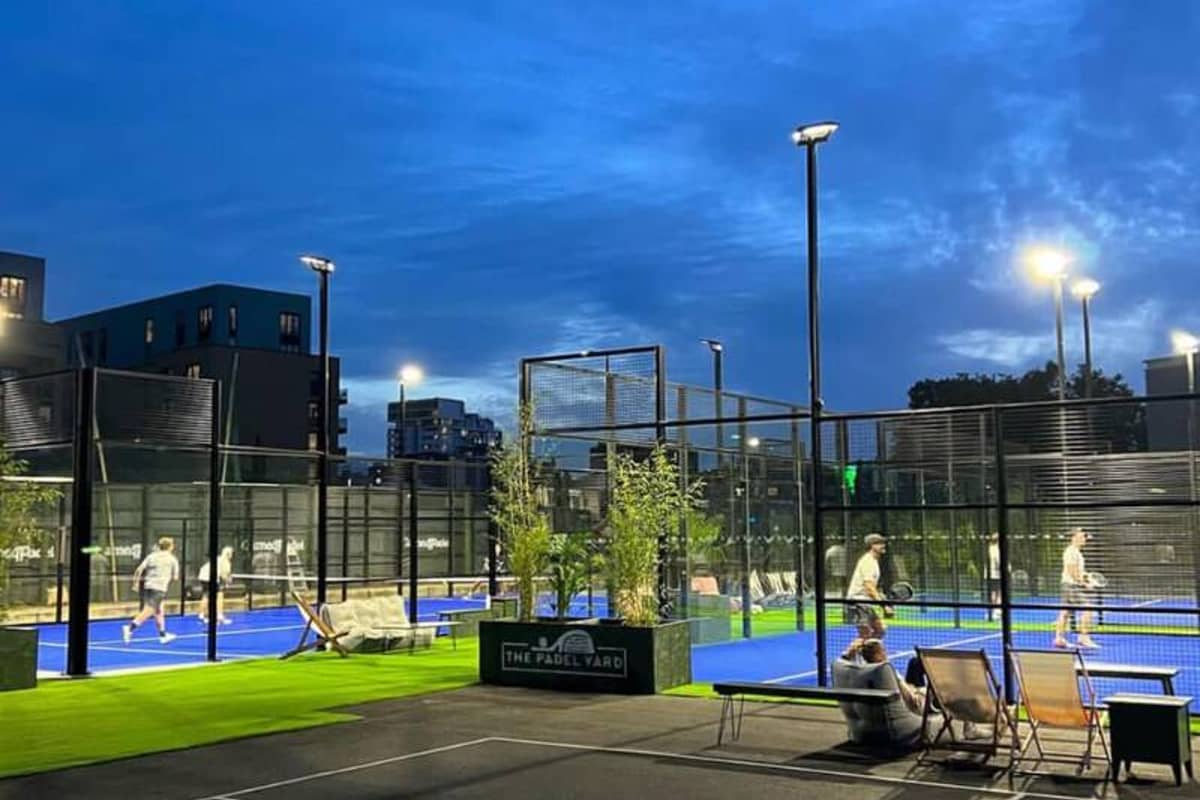 Evening view of The Padel Yard's outdoor courts with players and an urban backdrop, illuminated by lights.
