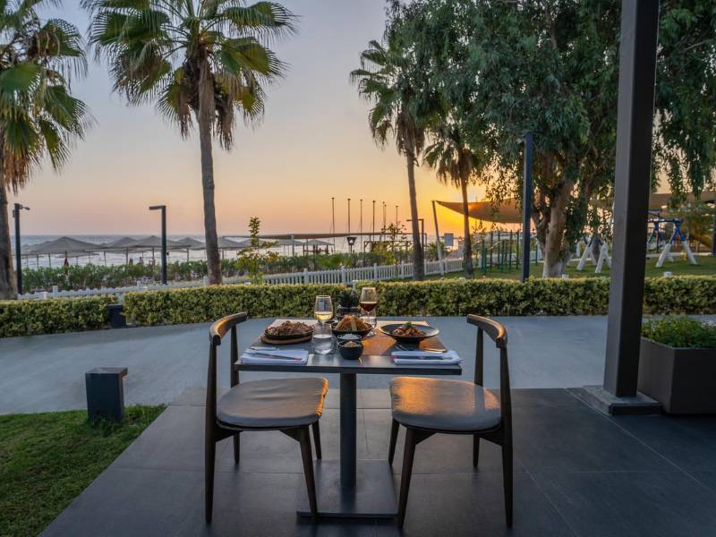 Outdoor dining setup at Paloma Family Holidays with a table for two, palm trees, and sunset in the background.