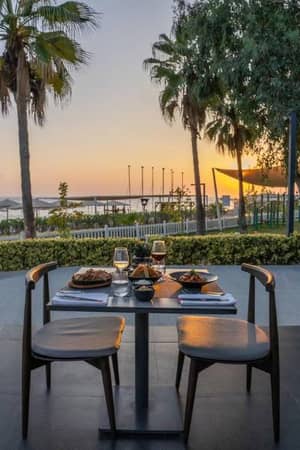 Outdoor dining setup at Paloma Family Holidays with a table for two, palm trees, and sunset in the background.