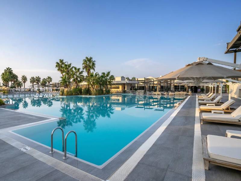 Pool view at Paloma Family Holidays, with sun loungers, umbrellas, and palm trees under a clear sky.