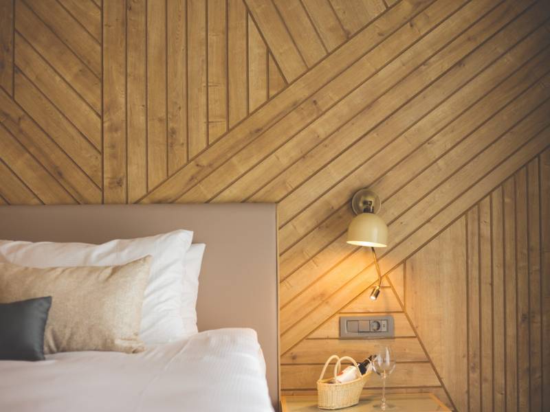 Stylish hotel room with a cosy bed, pillows, a wicker basket on a side table, and a diagonally patterned wooden wall with a wall lamp.