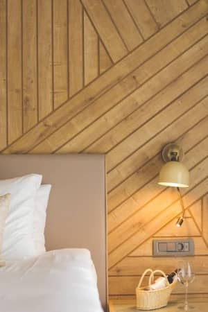Stylish hotel room with a cosy bed, pillows, a wicker basket on a side table, and a diagonally patterned wooden wall with a wall lamp.