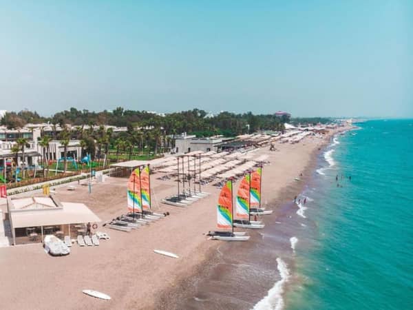 Beach at Paloma Gallery with colourful windsurfing sails, sandy shore, calm azure water, and parasol-covered lounge areas.