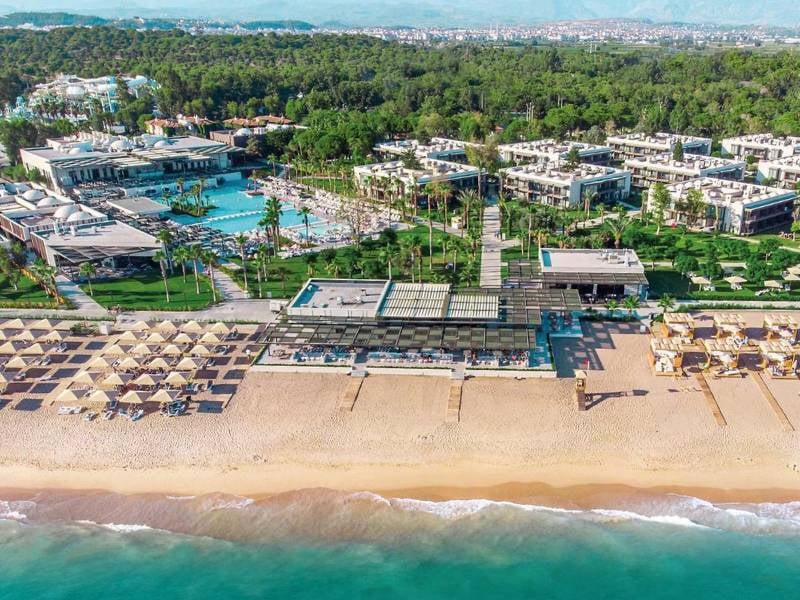 Aerial view of Paloma Gallery resort with a sandy beach, sun loungers, a swimming pool, and modern buildings set in lush greenery.