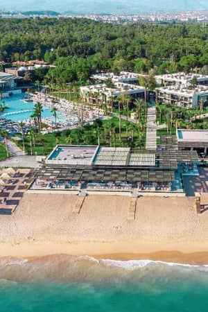 Aerial view of Paloma Gallery resort with a sandy beach, sun loungers, a swimming pool, and modern buildings set in lush greenery.