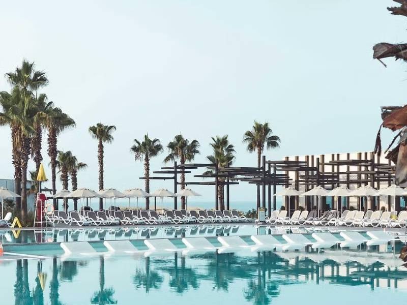 Luxurious pool area at Paloma Gallery with white sun loungers, umbrellas, and palm trees under a clear blue sky.