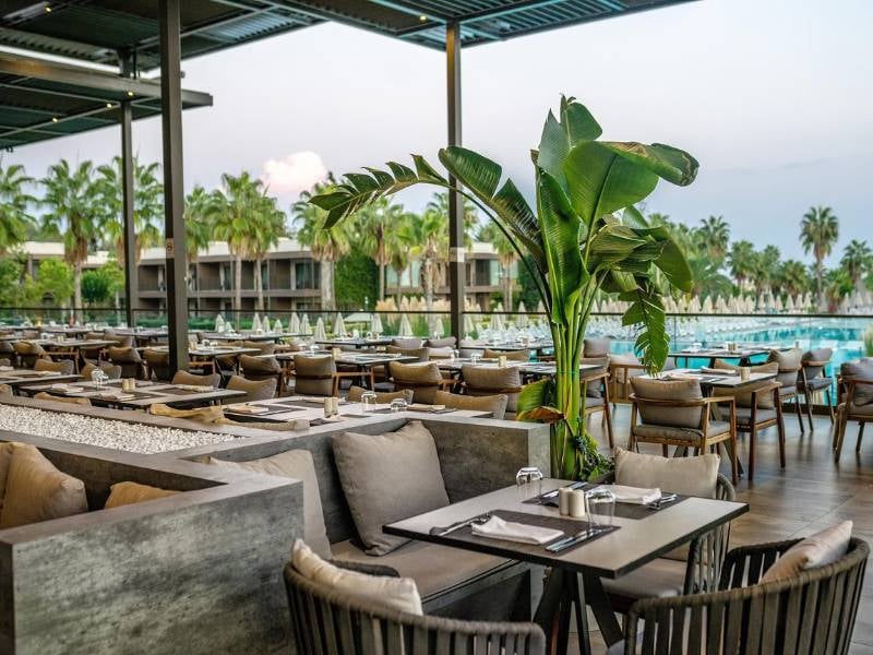 Luxurious outdoor dining area at Paloma Gallery - Restaurant with wooden tables, chairs, greenery, and a view of the pool.