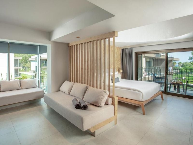 Modern hotel room at Paloma Gallery with a large bed, neutral sofa, wooden partition, and balcony overlooking gardens.