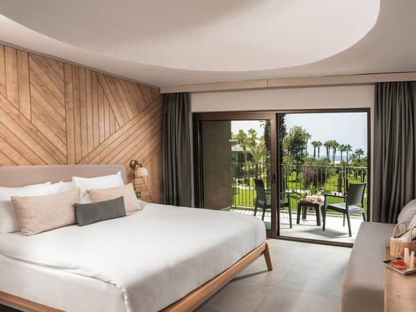 Hotel room at Paloma Orenda with a large bed, wooden decor, and a balcony offering a sea view and palm trees.