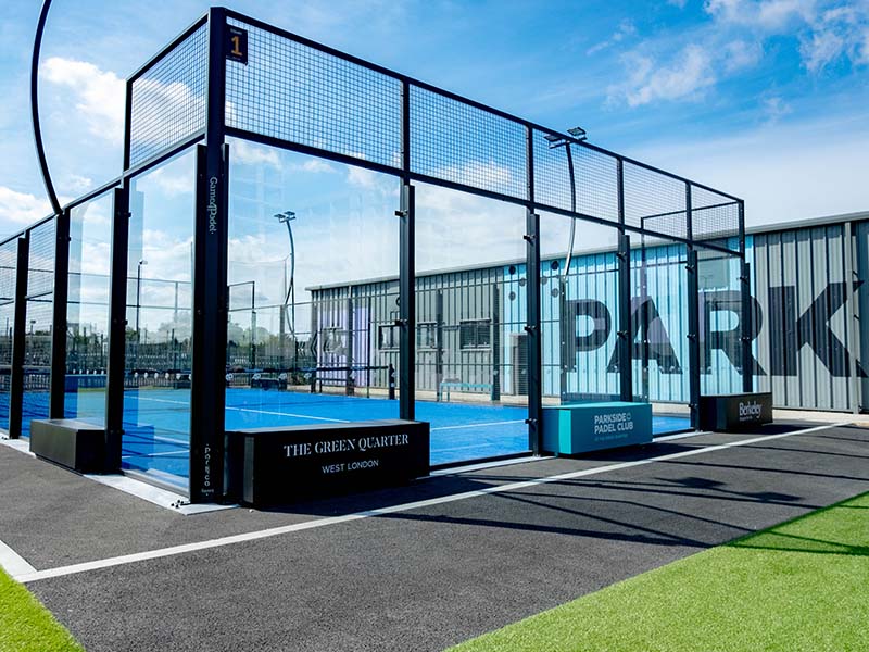 Outdoor Padel tennis court with blue surface and glass walls at Parkside Padel Club under a clear blue sky.