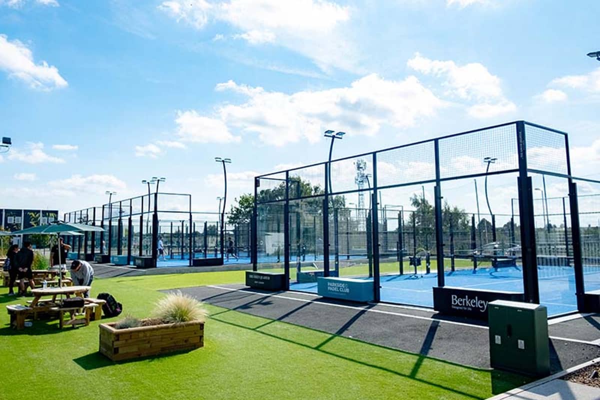 Outdoor padel tennis courts at Parkside Padel with players, surrounded by greenery and seating, on a sunny day.