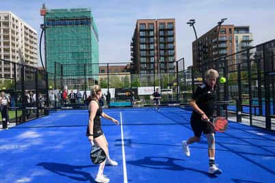 Two people playing padel on an outdoor court in Parkside, West London, with modern buildings in the background.