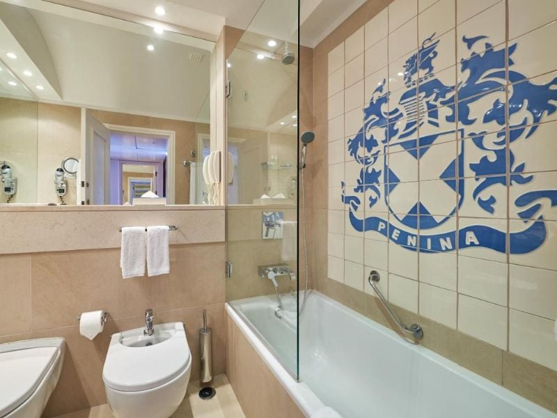 Modern bathroom at Penina Hotel with a bathtub, chrome fixtures, and the hotel's emblem on the tiled wall.