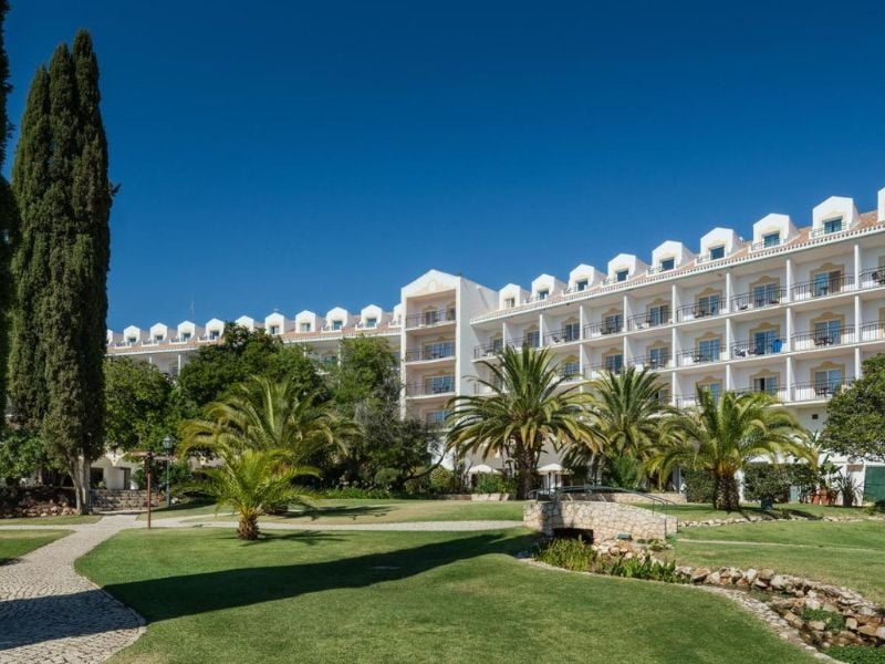 Exterior view of Penina Hotel with elegant multi-storey architecture, balconies, lush lawns, and palm trees under a clear blue sky.