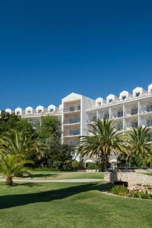 Exterior view of Penina Hotel with elegant multi-storey architecture, balconies, lush lawns, and palm trees under a clear blue sky.