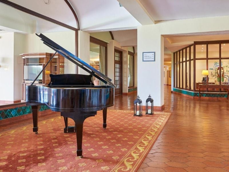 Penina Hotel lobby with a grand piano on a red carpet, surrounded by wooden decor and large windows.