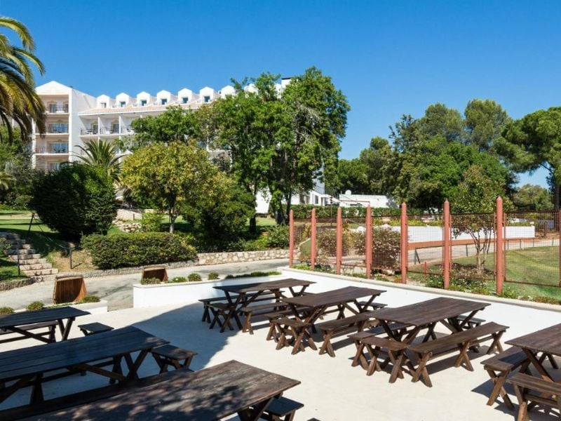 Outdoor lounge area at Penina Hotel with wooden picnic tables, trees, and tennis courts in the background.