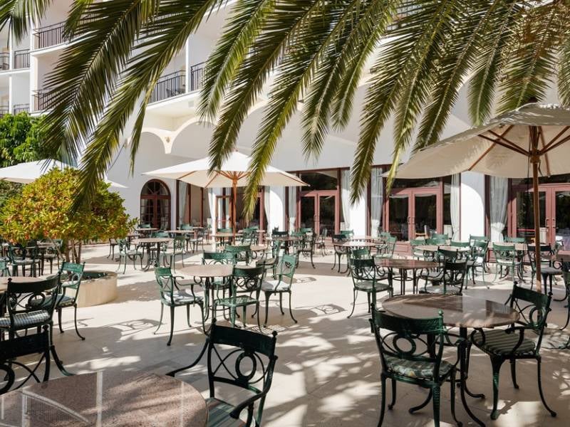 Outdoor dining area at Penina Hotel with green wrought iron tables and chairs under umbrellas and palm trees against the hotel's white facade.