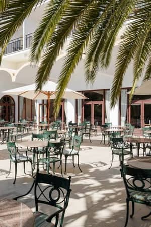 Outdoor dining area at Penina Hotel with green wrought iron tables and chairs under umbrellas and palm trees against the hotel's white facade.