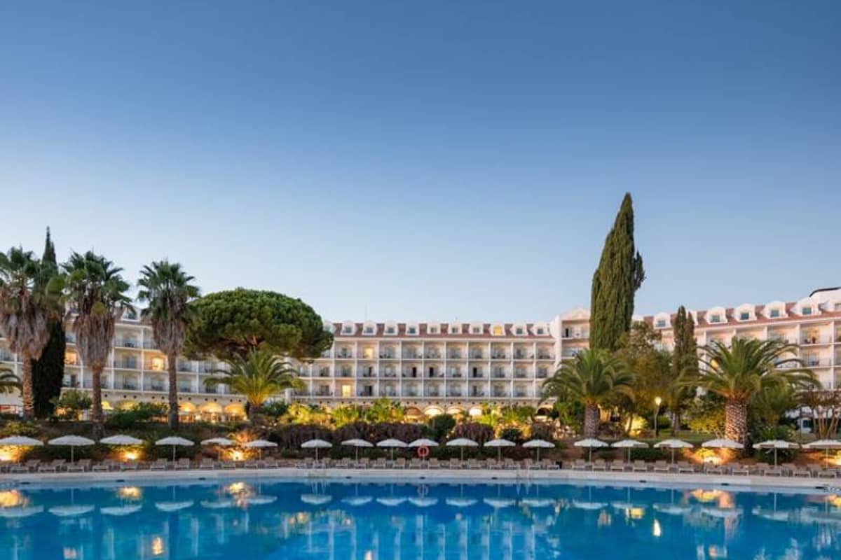 Panoramic view of Penina Hotel's façade, lush palm trees, and large swimming pool under a clear sky.