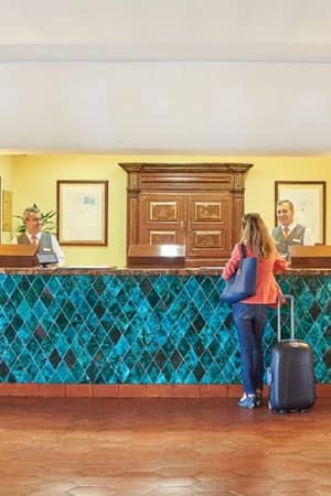 Reception area of the Penina Hotel with a turquoise tiled desk, two receptionists, and a female guest wheeling a suitcase.