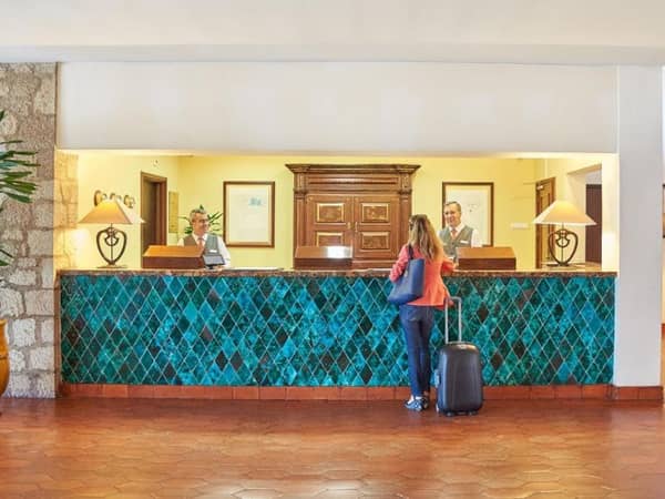 Reception area of the Penina Hotel with a turquoise tiled desk, two receptionists, and a female guest wheeling a suitcase.