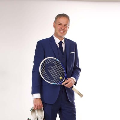 Peter Jones in a blue suit holding a tennis racket and white trainers, highlighting his dual interest in business and sports.