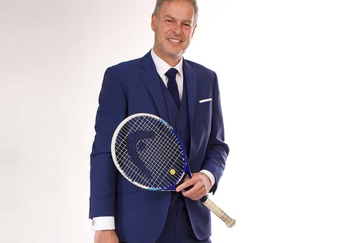 Peter Jones in a blue suit holding a tennis racket and white trainers, highlighting his dual interest in business and sports.