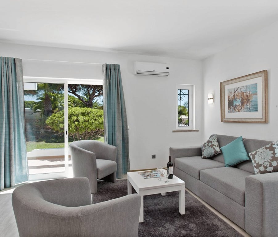 Living area of a Prado Villas 2-bedroom deluxe villa with grey sofas, a white coffee table, armchairs, and garden access through glass doors.