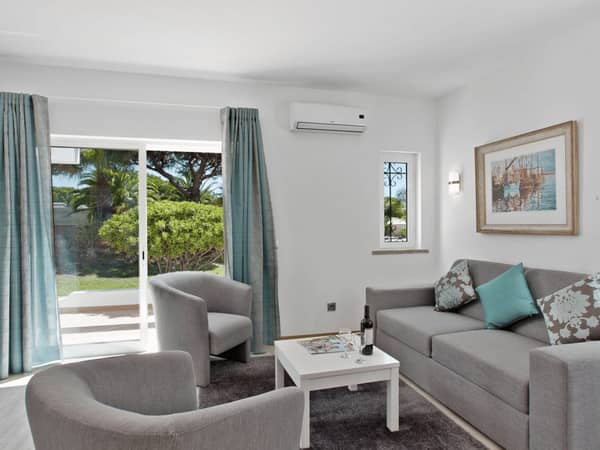Living area of a Prado Villas 2-bedroom deluxe villa with grey sofas, a white coffee table, armchairs, and garden access through glass doors.