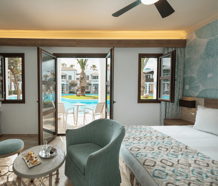 Interior of the Royal Mare Bungalow Sharing Pool with a bed, seating area, and windows opening to a shared pool area.