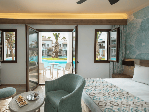 Interior of the Royal Mare Bungalow Sharing Pool with a bed, seating area, and windows opening to a shared pool area.