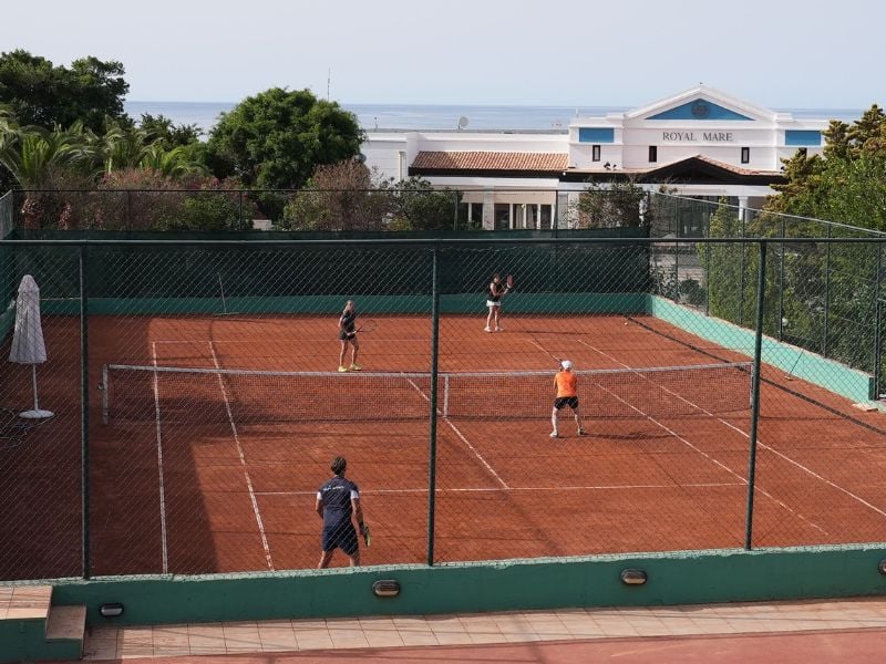 Players on a clay tennis court at Royal Mare hotel with lush greenery and hotel building in the background.