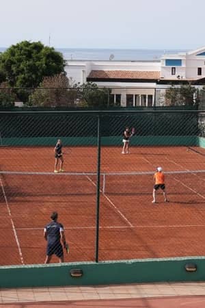 Players on a clay tennis court at Royal Mare hotel with lush greenery and hotel building in the background.