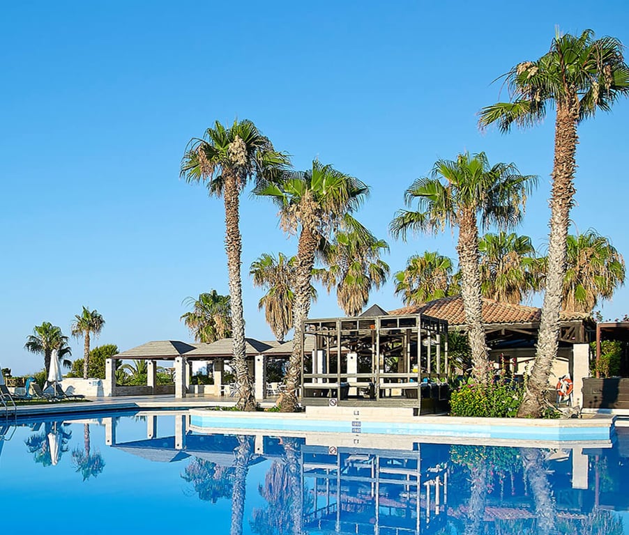Royal Mare setting with a blue swimming pool, surrounded by palm trees and a wooden structure under a clear sky.