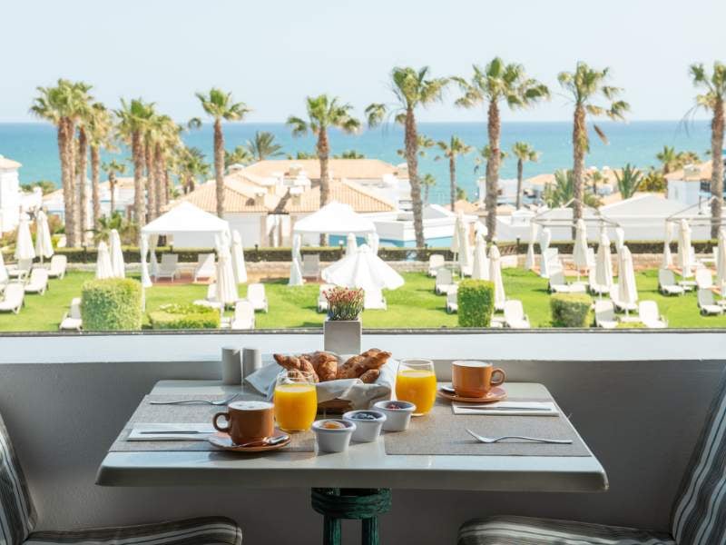 Breakfast table with croissants, orange juice, and coffee, overlooking a garden with palm trees and sea view at Royal Mare.