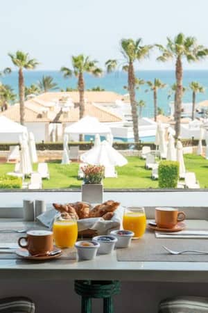 Breakfast table with croissants, orange juice, and coffee, overlooking a garden with palm trees and sea view at Royal Mare.