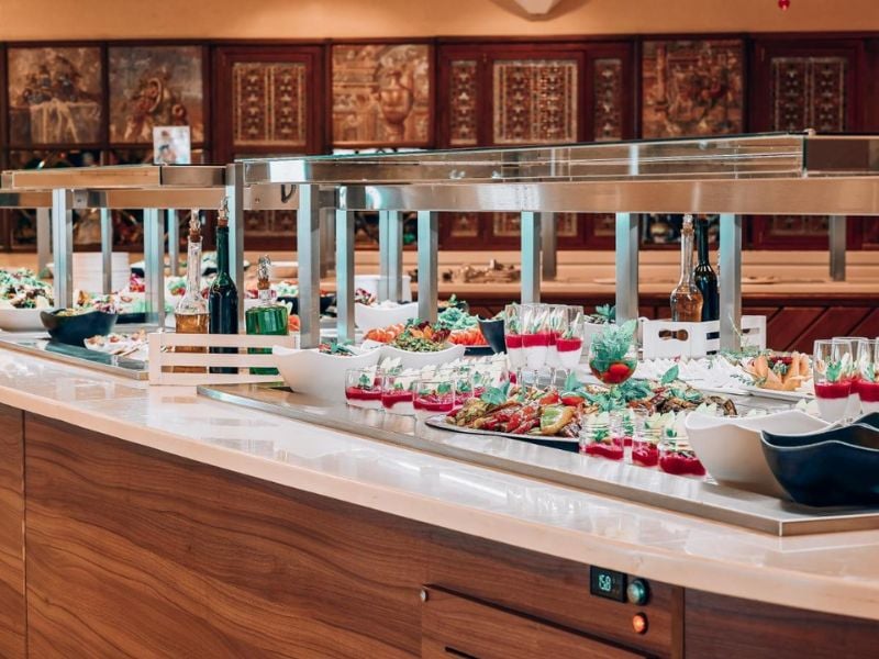 Luxurious buffet at Royal Mare Gallery with gourmet dishes, salads, drinks, and elegant presentation on a wooden counter with stainless steel warming lamps.