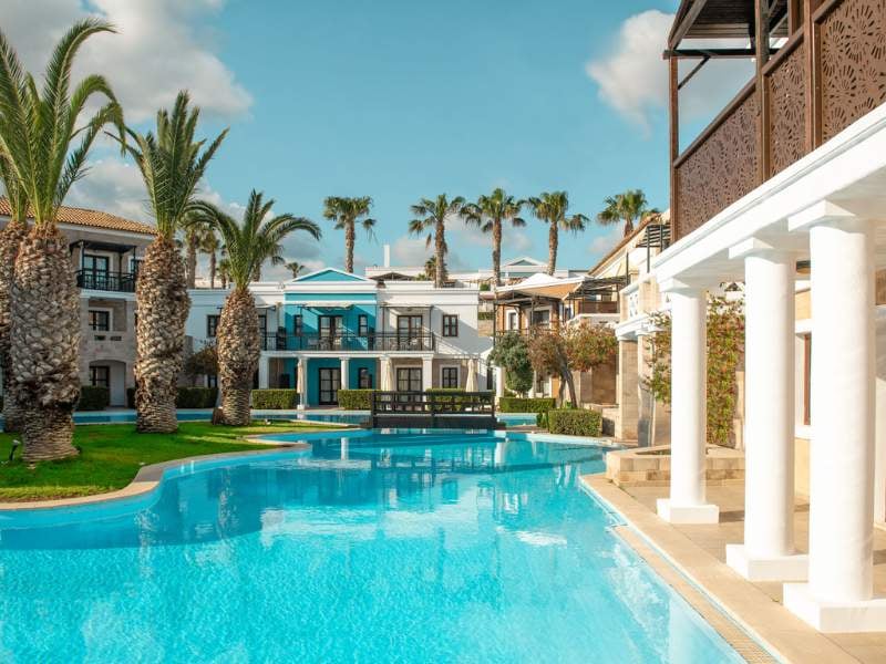 Luxurious pool at Royal Mare with palm trees and Mediterranean-style villas in the background, under a clear blue sky.