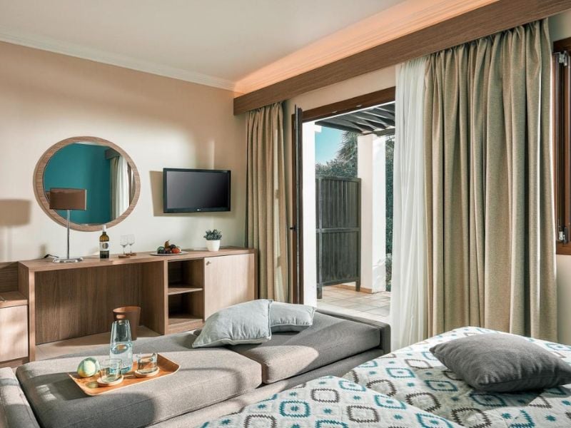Modern hotel room at Royal Mare with a bed, patterned bedding, wooden console, round mirror, flat-screen TV, and balcony door open to natural light.