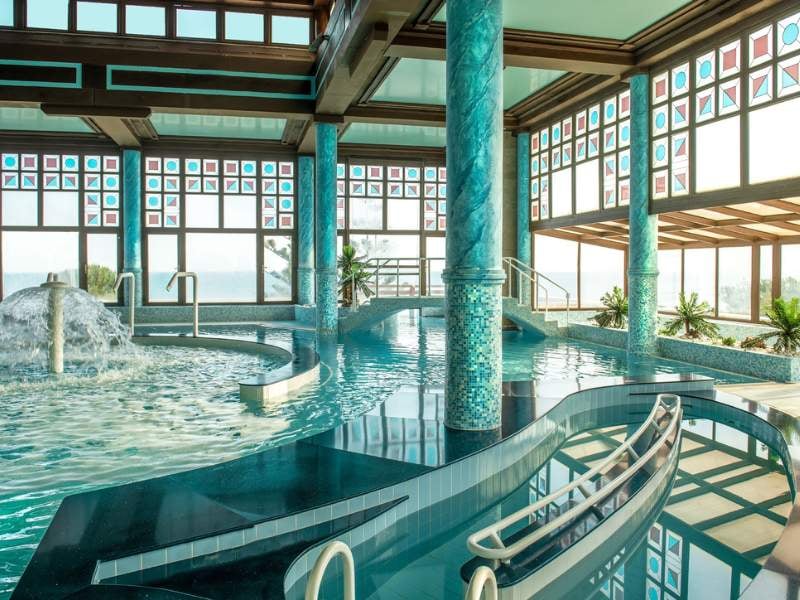 Luxurious indoor spa area at Royal Mare Gallery, featuring a large pool with mosaic columns and panoramic windows with views outside.