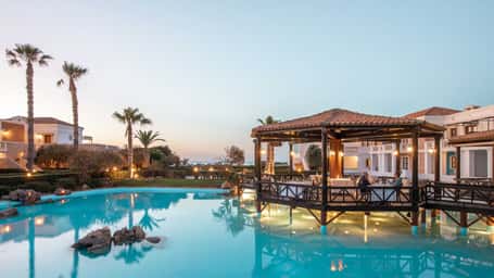 Royal Mare resort's poolside pavilion surrounded by palm trees and white buildings with an evening sky and ocean view.
