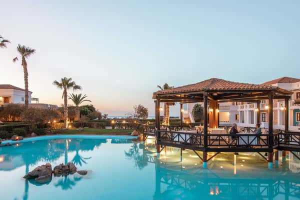 Royal Mare resort's poolside pavilion surrounded by palm trees and white buildings with an evening sky and ocean view.