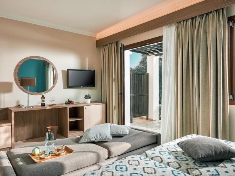 Luxurious hotel room at Royal Mare with a grey chaise lounge, flat-screen TV, round mirror, and access to private terrace through sliding doors.